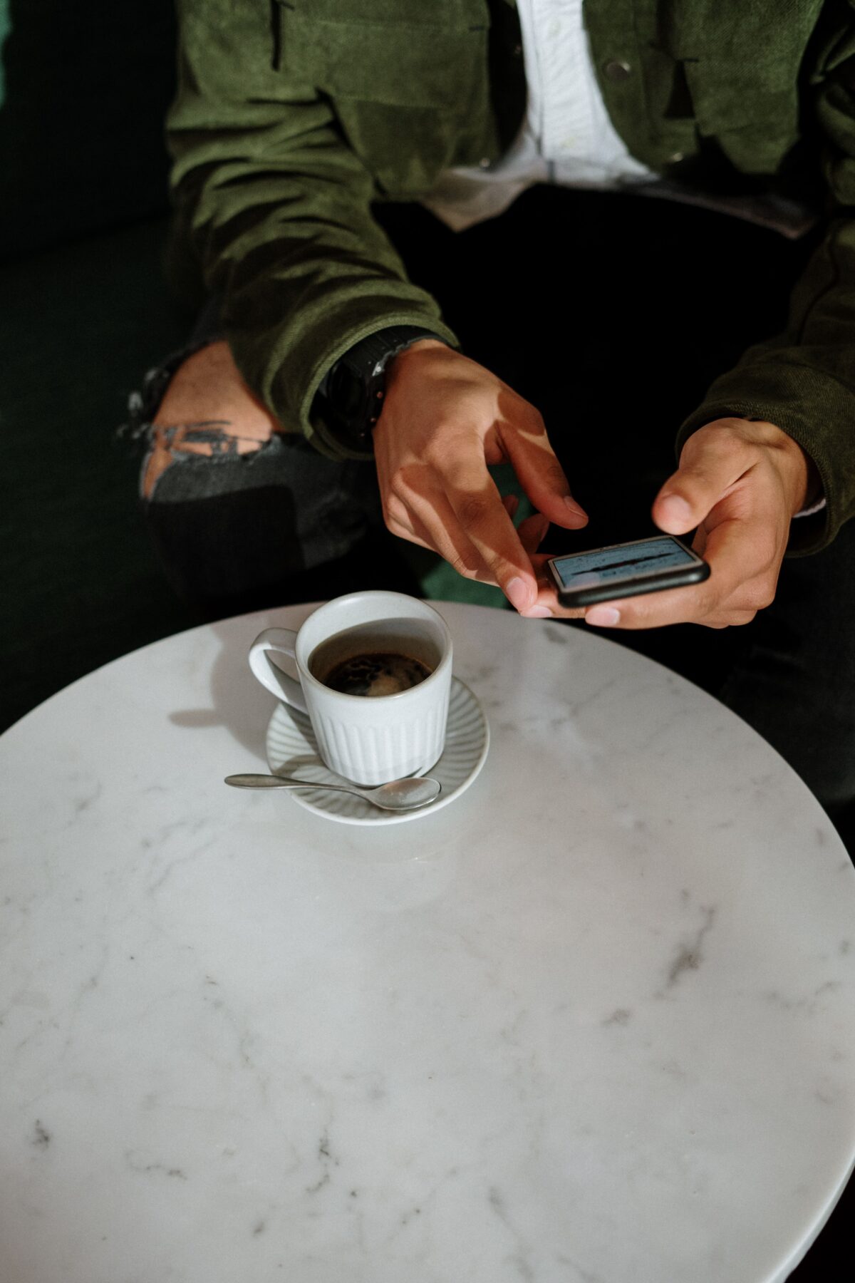 A person wearing a green jacket and ripped jeans sits at a round white marble table with a cup of coffee, holding and using a smartphone with both hands.